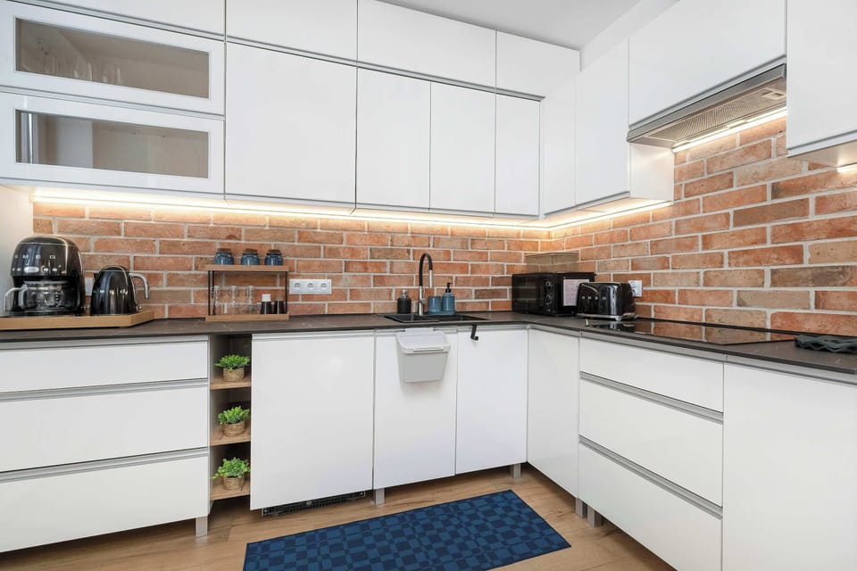 A modern L-shaped kitchen showcases white cabinetry paired with a detailed brick backsplash and under-cabinet lighting. Black appliances and a dark countertop create a sharp, stylish contrast.