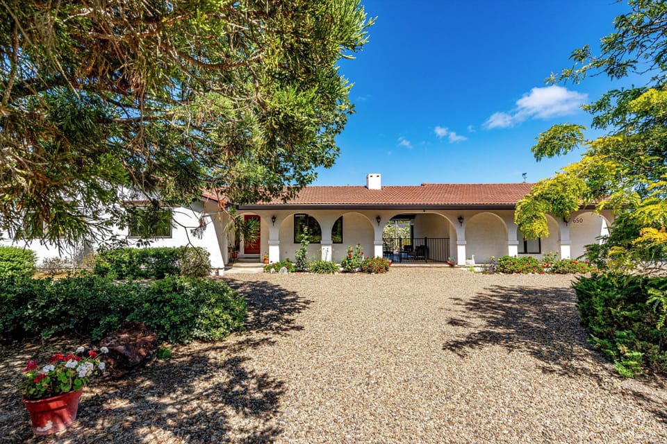White stucco property with Spanish-style arched portico, red tile roof, and mature trees framing the gravel driveway entrance.