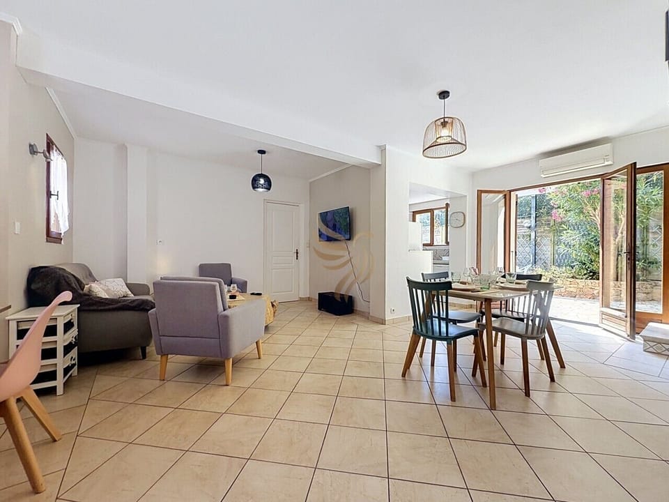 Un grand salon lumineux avec coin repas et portes-fenêtres donnant sur une terrasse arborée. Canapé, fauteuils et table dressée pour 4 personnes.