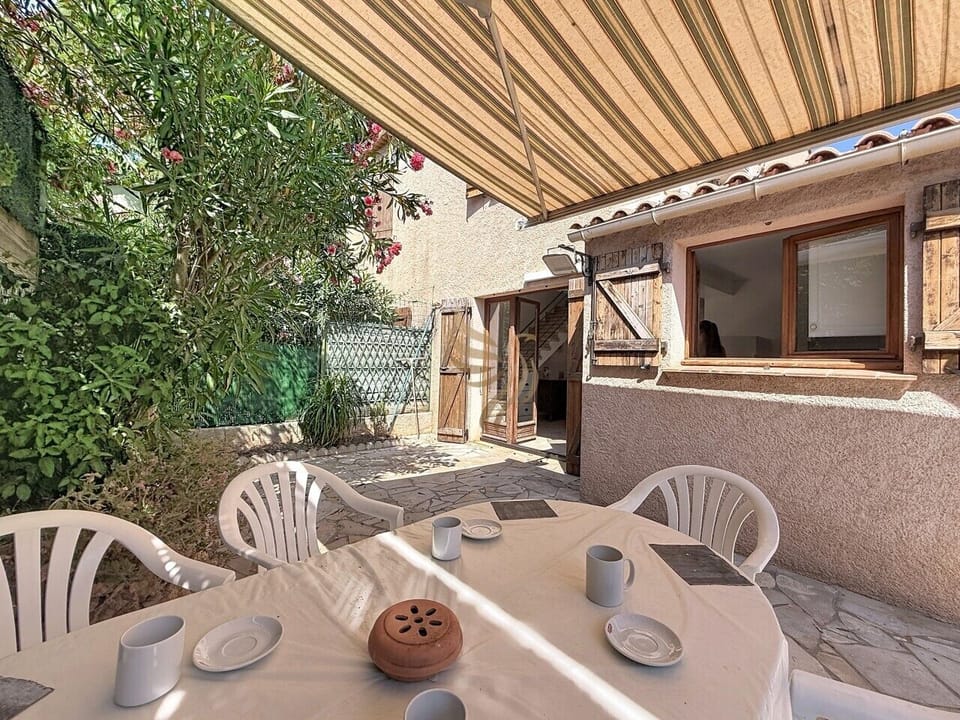 Un angle légèrement plus large de l`espace repas sur la terrasse, montrant la table dressée, le store déplié protégeant du soleil, ainsi que la façade en crépi avec ses ouvertures en bois et la végétation environnante.