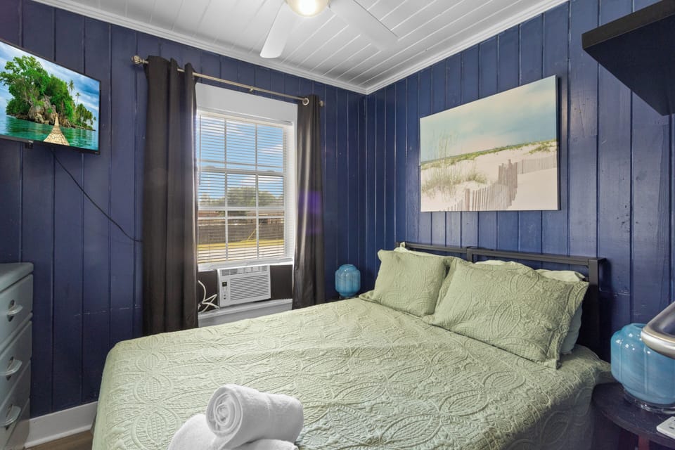 The primary bedroom offers a wall-mounted flat-screen TV, refreshing ceiling fan, and beautiful coastal artwork above the bed.
