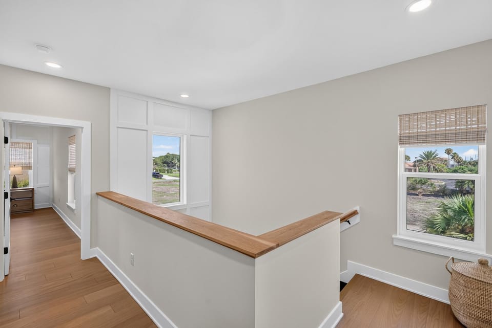 Upstairs landing with light oak railing, beige walls, bamboo shades, and views of palm trees outside two windows.