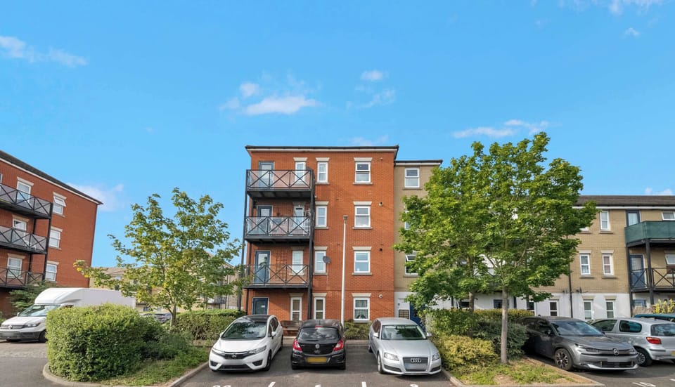 This brick apartment building features multi-level private balconies and a paved residential parking lot surrounded by green trees.