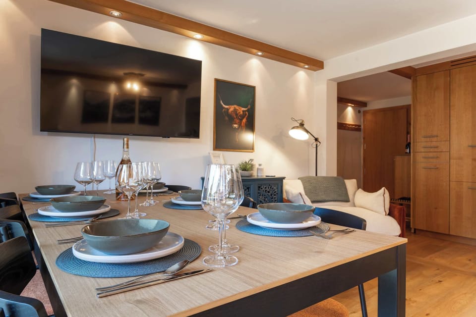 Gather around this beautifully set dining table, featuring elegant place settings and glassware, set against a warm living area complete with a wall-mounted TV and a cozy armchair.