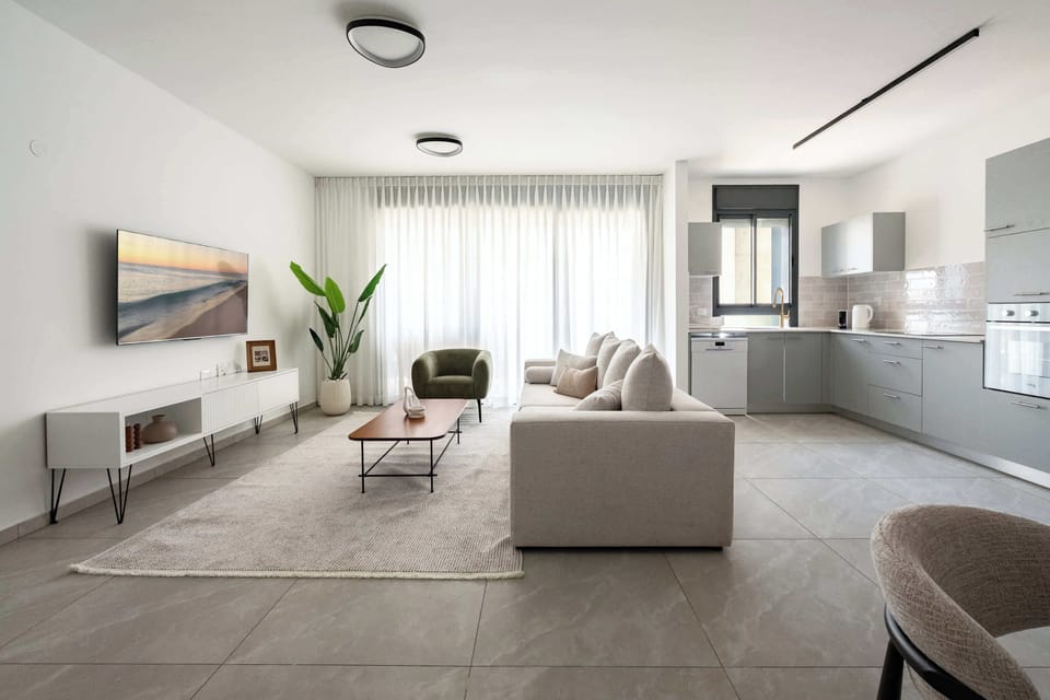 Contemporary living room with mounted television, plush sofa, botanical accents, and visible kitchenette beyond.
