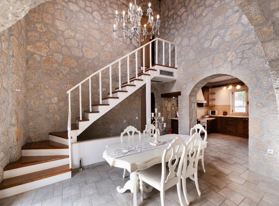Stone dining room with a white table set beneath a staircase and crystal chandelier.