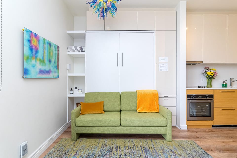 Green fabric sofa with orange accents sits on a patterned rug against white built-in cabinetry and colorful wall art.
