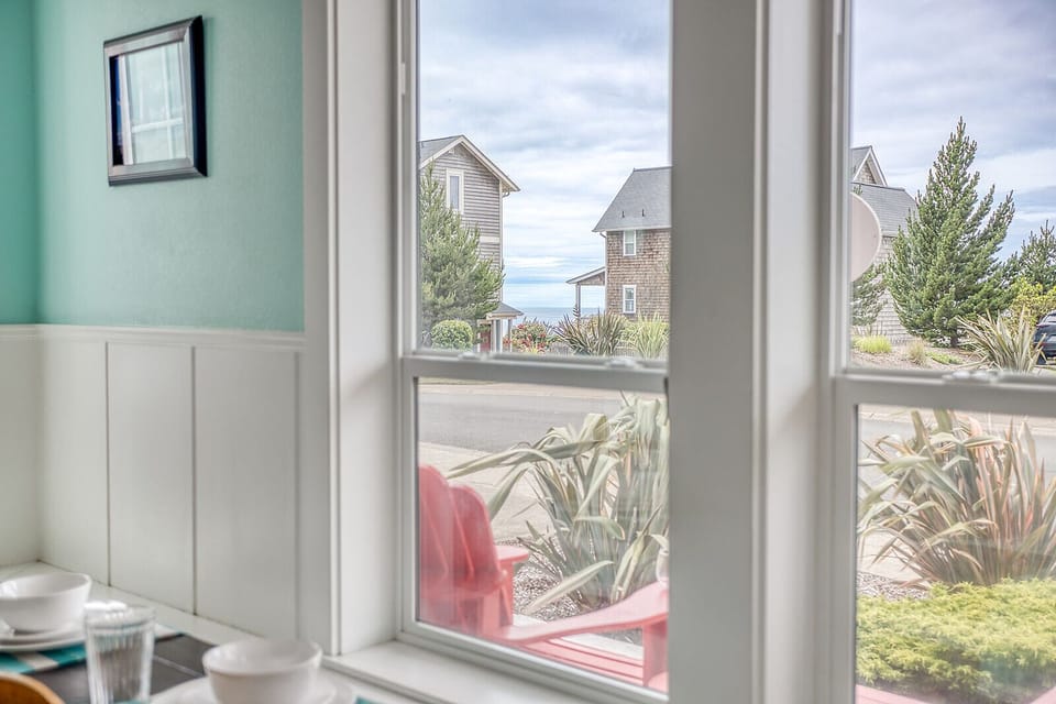 A peaceful coastal neighborhood frames the view through bright windows, with nearby residential homes and ocean glimpses visible beyond the street. Local greenery and seaside charm characterize the surrounding area.