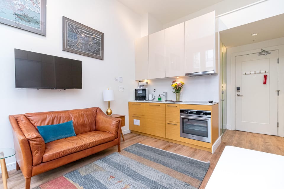 Living area with caramel leather sofa, blue accent pillow, wall-mounted TV, and compact kitchen featuring light wood cabinets.