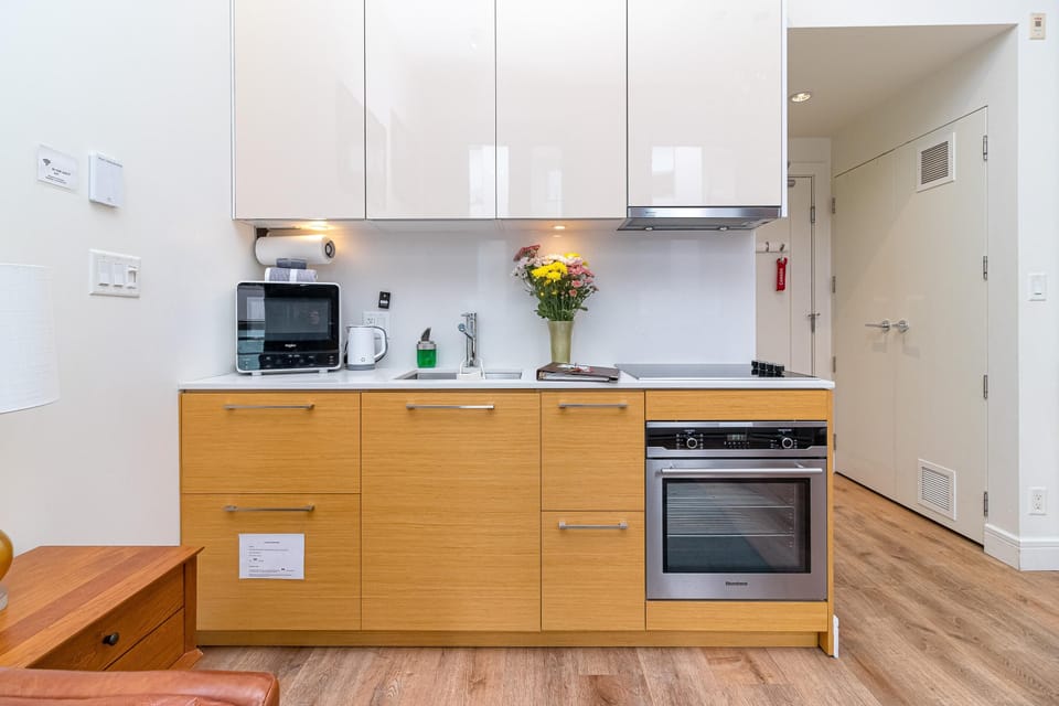 Kitchen area featuring light wood lower cabinets, white high-gloss upper cabinets, stainless steel oven, and under-cabinet lighting.