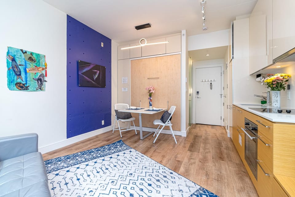 Dining table with white chairs set against a wood-paneled wall and blue accent panel in the studio living area.