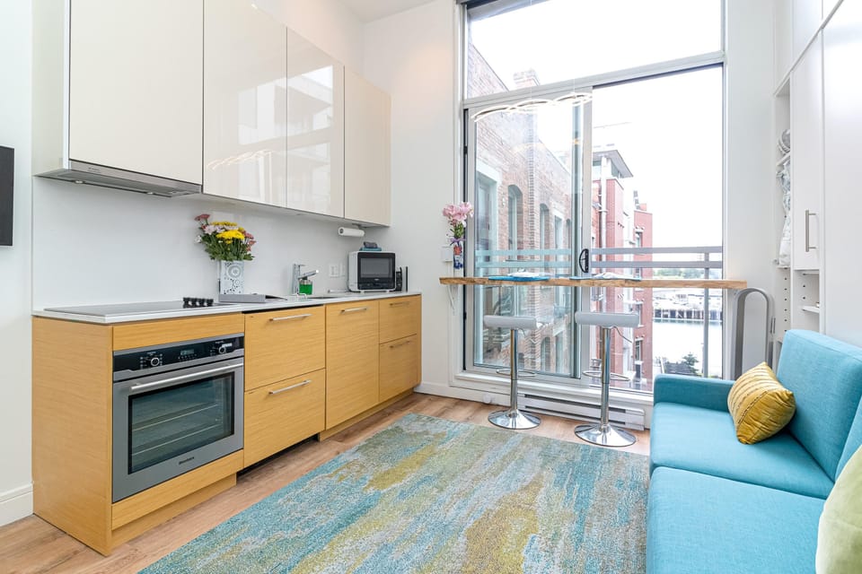 Compact kitchen features wood lower cabinets, glossy upper storage, and a built-in oven next to floor-to-ceiling windows.