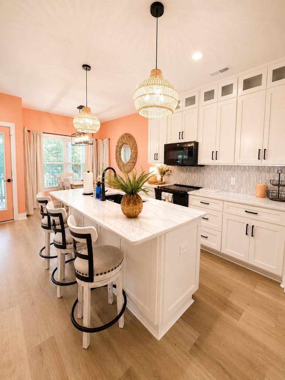Three woven pendant lights hang above a white kitchen island with beige cushioned bar stools and wood flooring.