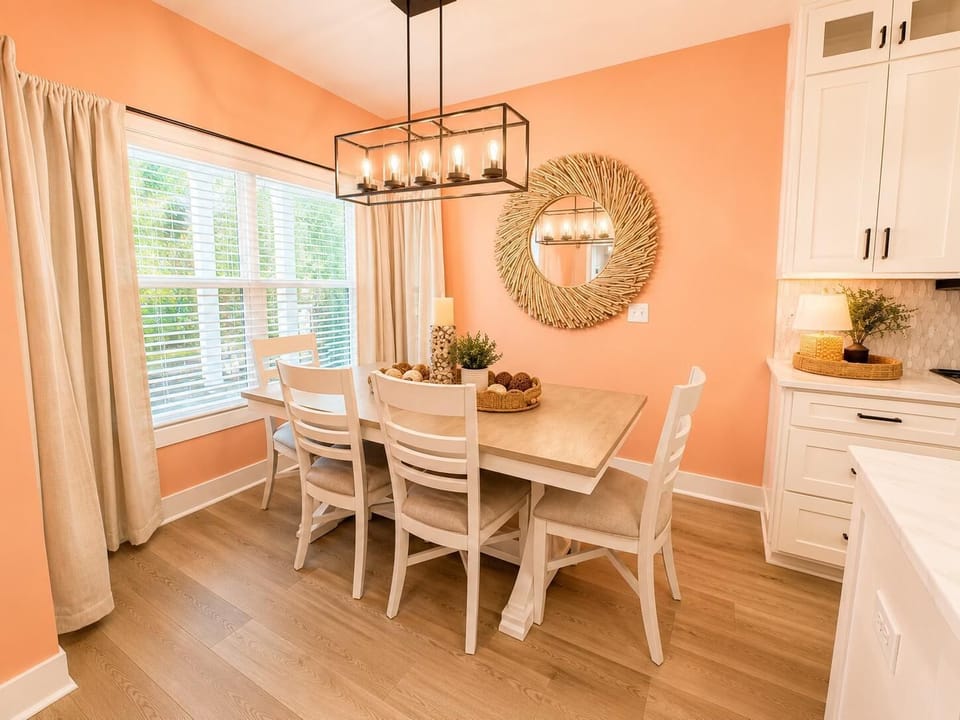 Light wood dining table with six cushioned white chairs under a black metal pendant light and coral walls.