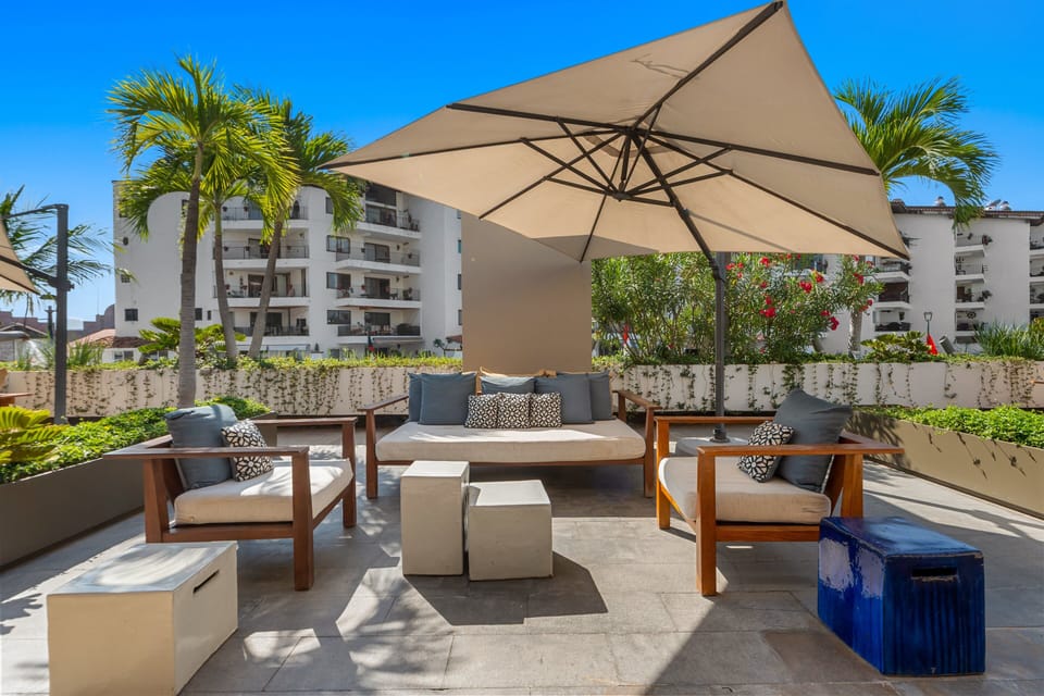 Lounge comfortably under a massive patio umbrella in stylish wood frame seating. Surrounded by lush planters, blooming flowers, and vibrant tropical palm trees.