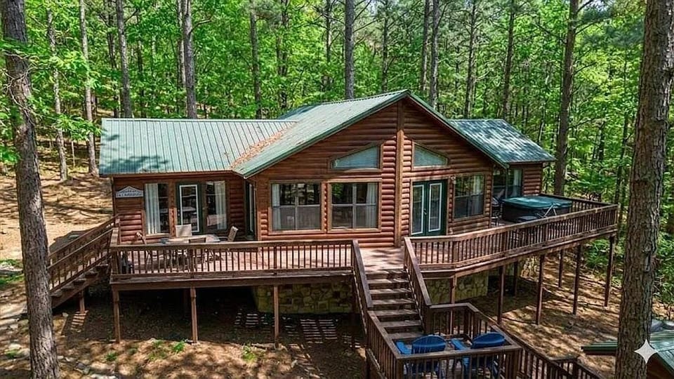 Aerial view of a log cabin rental nestled among lush forest, featuring a wraparound deck and green metal roof in a tranquil woodland setting.