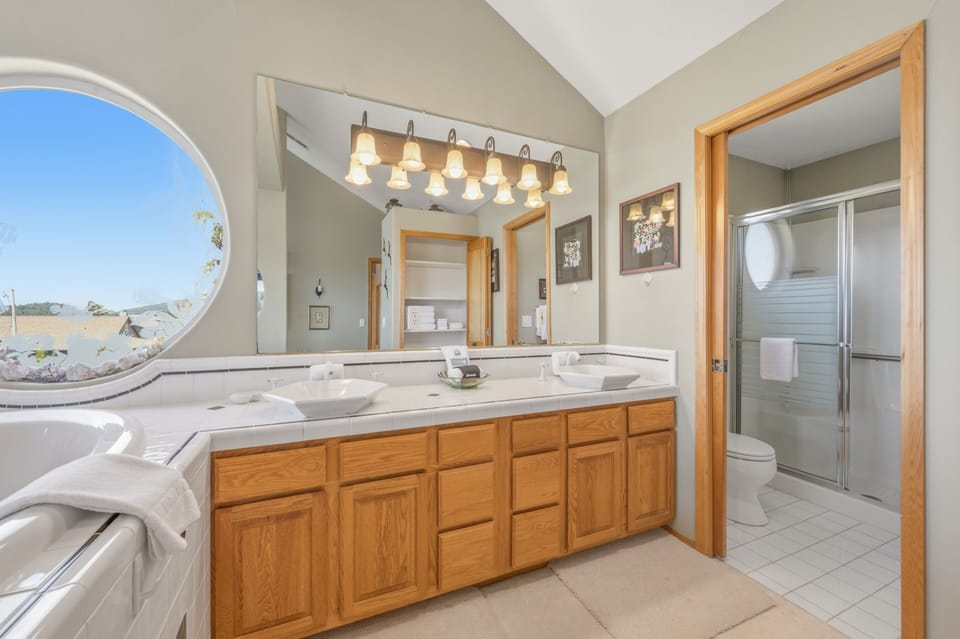 Enjoy your morning routine in this spacious bathroom with dual sinks, warm wood cabinetry, and abundant natural light streaming through the unique circular window overlooking scenic views.