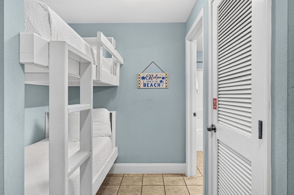 Your beach-themed bunk room features coastal blue walls and crisp white linens, perfect for restful nights after sunny days. Note the charming beach sign welcoming you to pure relaxation.