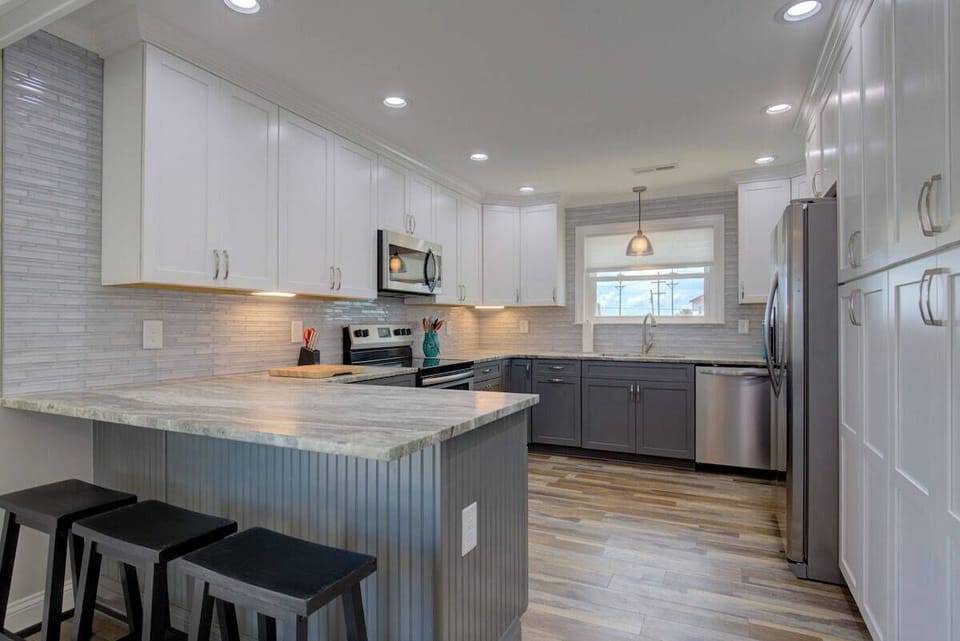 A modern kitchen with white cabinets, a central island, bar stools, stainless steel appliances, and a window providing natural light.
