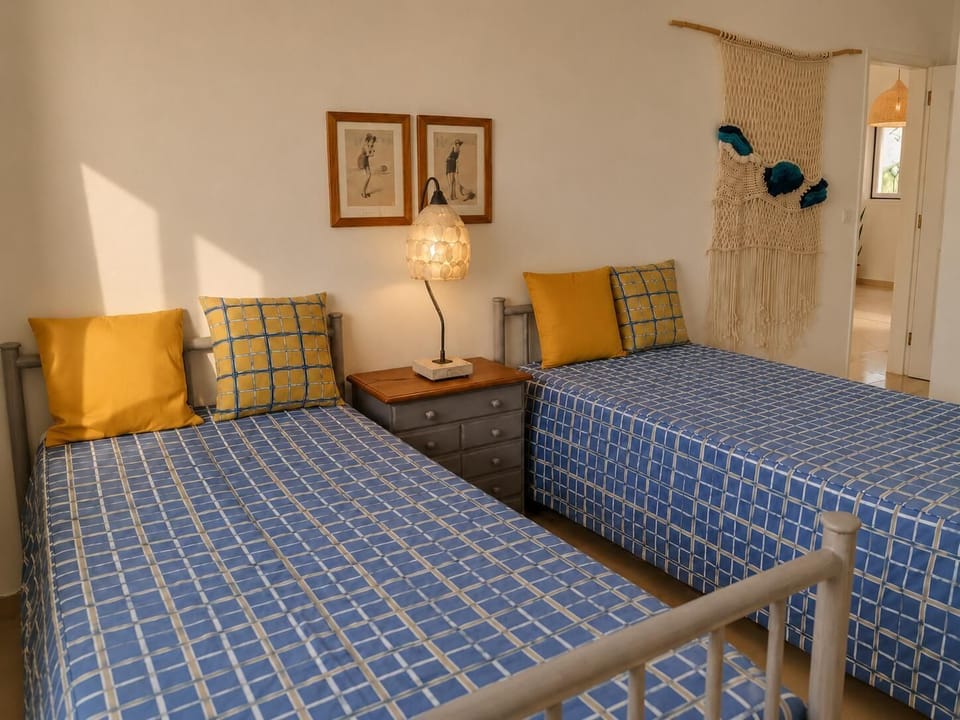 An alternate perspective of the twin bedroom, showcasing the blue-and-yellow plaid bedding, solid yellow pillows, a central grey nightstand with a lamp, and a macrame tapestry near the open doorway.