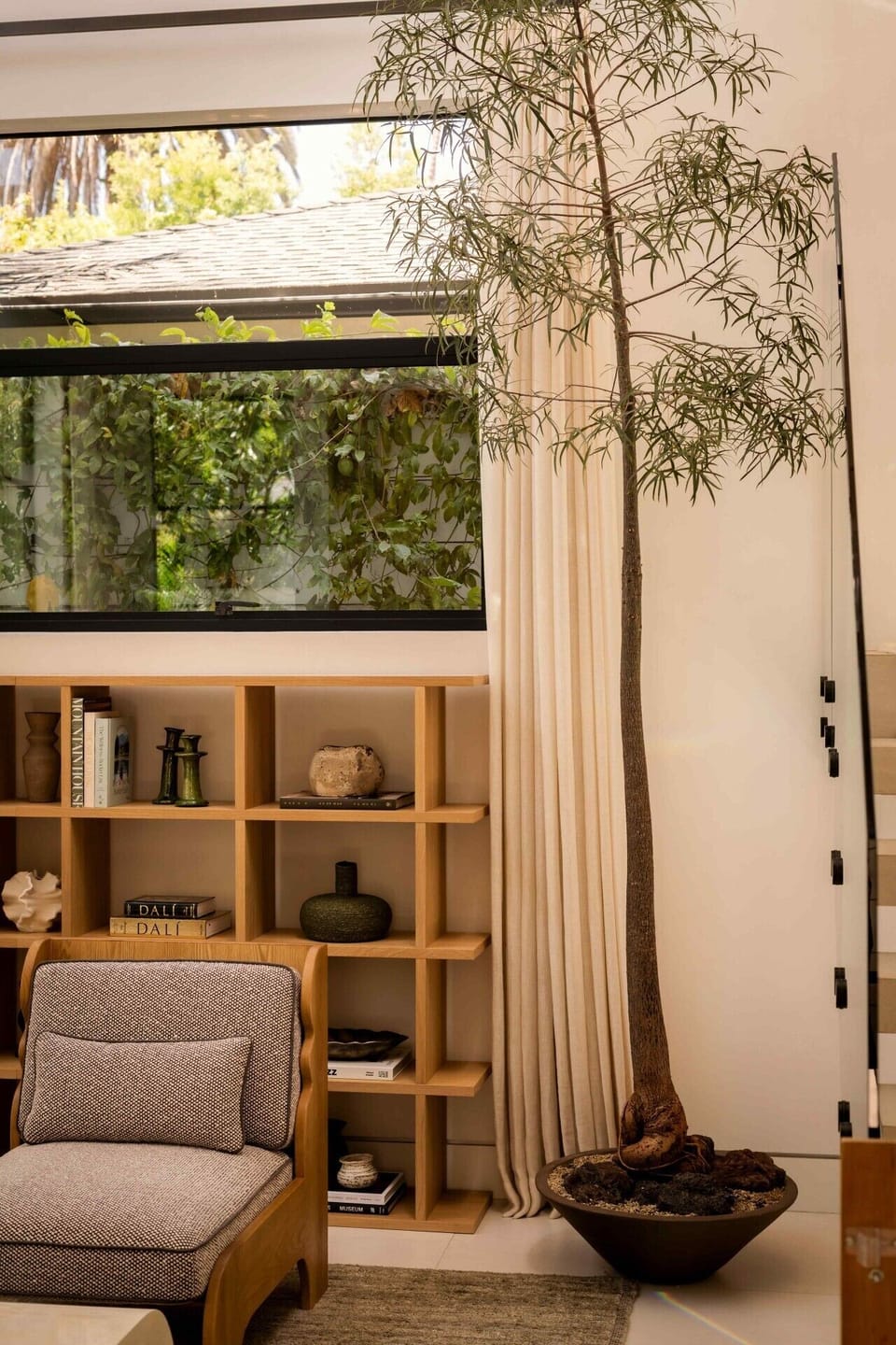 Tucked beneath a picture window overlooking swaying palms and greenery, this cozy corner invites you to slow down. A sculptural lounge chair pairs with an open-shelf display of art books and curated ceramics, while a potted tree and soft linen curtains ad