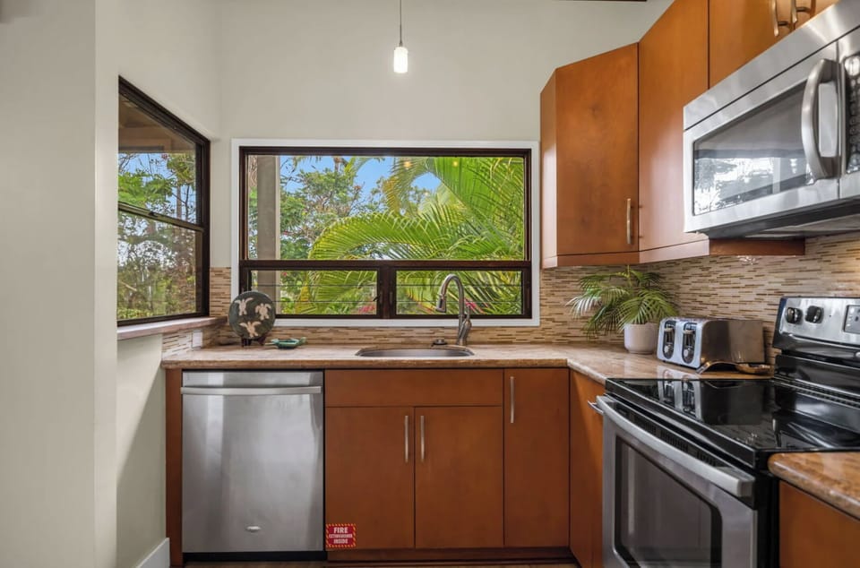 Unit 1: Prepare meals in a bright kitchen featuring stainless steel appliances, rich wood cabinetry, and a large window framing tropical views.