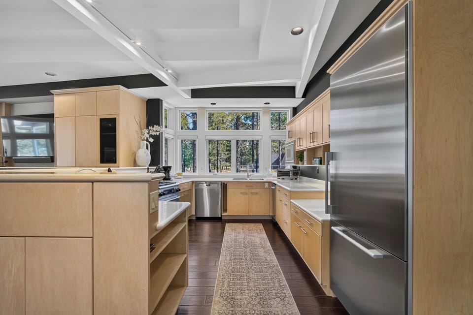 Your chef's kitchen features warm wood cabinetry, professional-grade stainless appliances, and abundant natural light streaming through floor-to-ceiling windows overlooking a peaceful wooded setting.