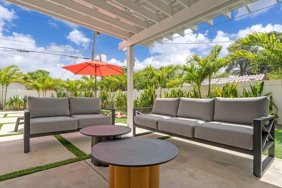 Shaded outdoor lounge with plush sofa seating and modern coffee tables under a pergola. A comfortable spot to relax poolside, surrounded by tropical greenery.