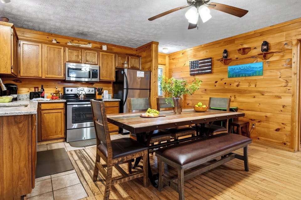 Dining area and kitchen featuring a rustic wooden table with bench seating and a ceiling fan.