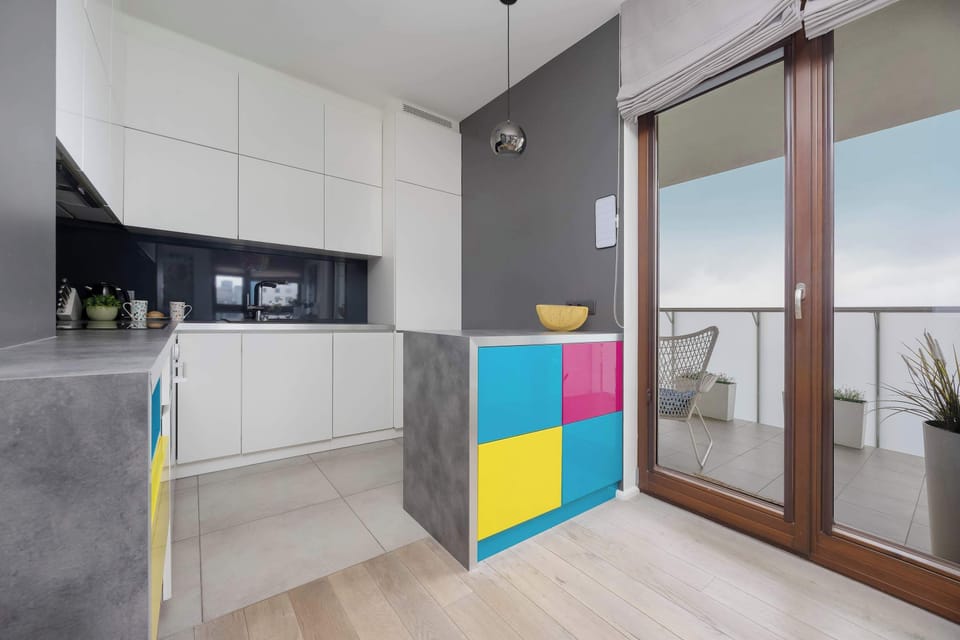 A modern kitchenette with colorful cabinets and plenty of space for preparing meals. Large balcony doors bring in plenty of natural light.