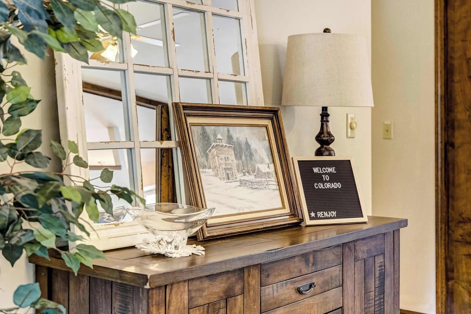 A rustic wood console table greets you just inside, styled with framed art, a decorative mirror, and warm lighting beneath an arched, multi-pane window that fills the entry with natural light.

