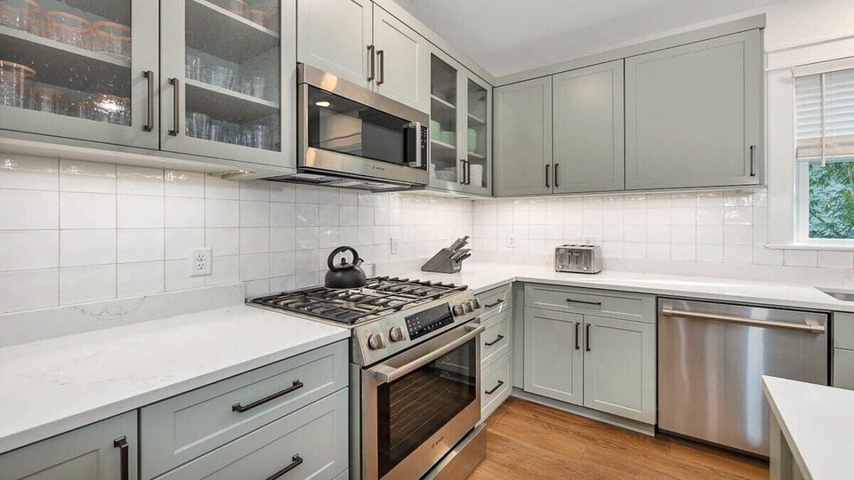 Your beautifully renovated kitchen awaits with soft sage cabinetry, gleaming white countertops, and modern stainless appliances—perfect for preparing morning coffee or homecooked meals.
