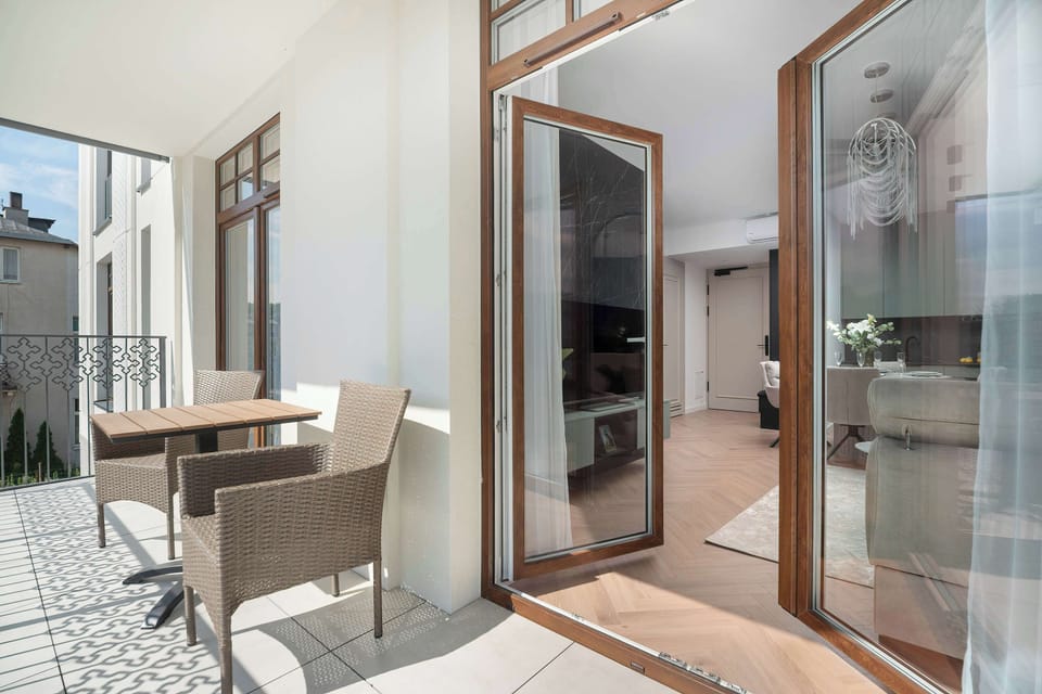 Open wooden balcony doors lead onto a tiled terrace furnished with two woven chairs and a coffee table. The view extends into the living room.