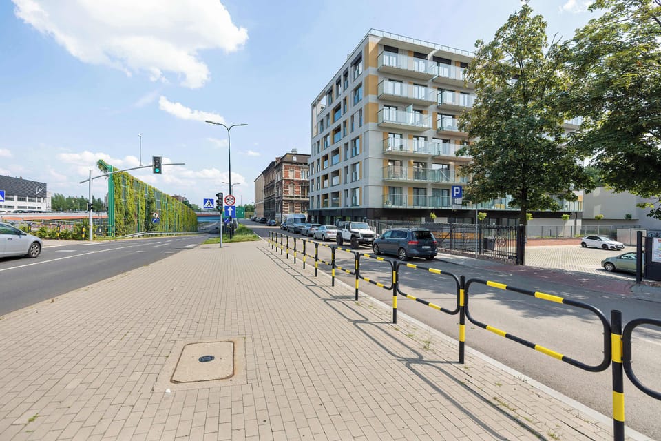 A street-level perspective showcases a wide paved sidewalk and crosswalk next to modern apartment buildings and a green noise-barrier wall.