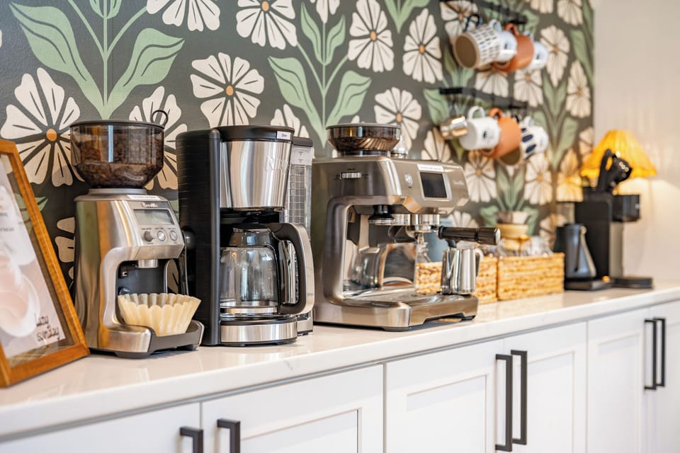 Exquisite coffee bar — Breville grinder, espresso machine, drip brewer, and mug rail against bold floral wallpaper. Morning sorted.