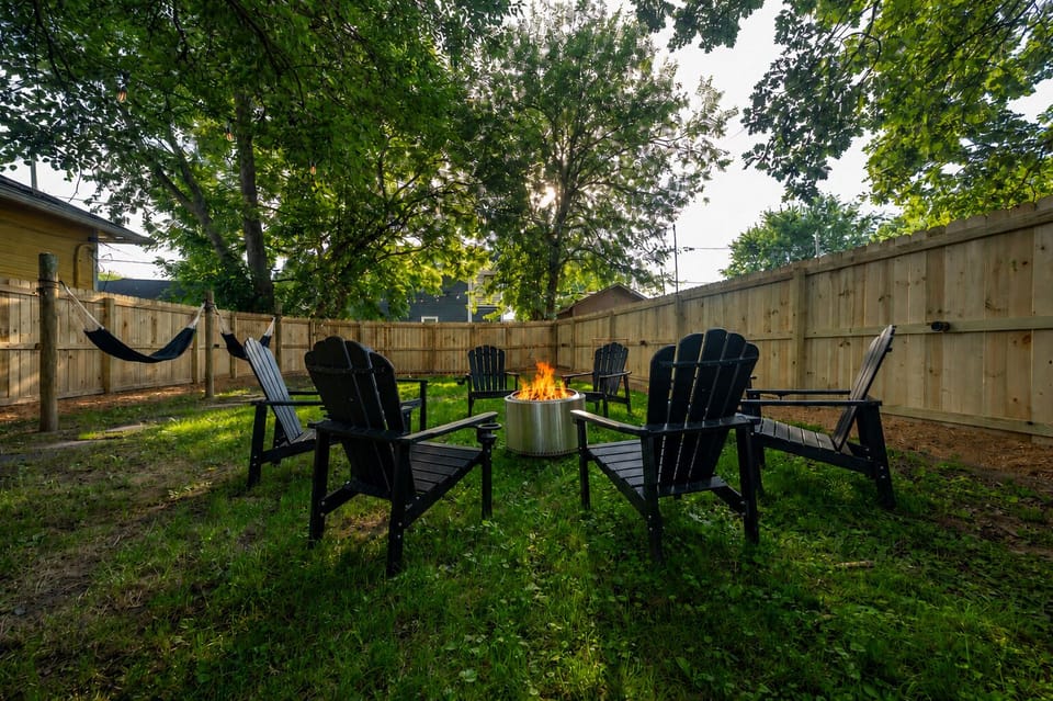 Fenced backyard with Adirondack chairs circling a lit solo stove, hammock, and mature shade trees — no one's going inside yet.