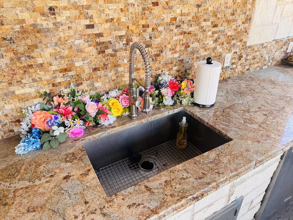 Outdoor kitchen sink with granite counters, stone backsplash, and prep space.
