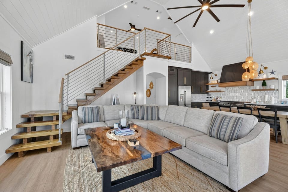 Coastal living room with sectional sofa, wood coffee table, staircase, kitchen view, and vaulted ceiling.
