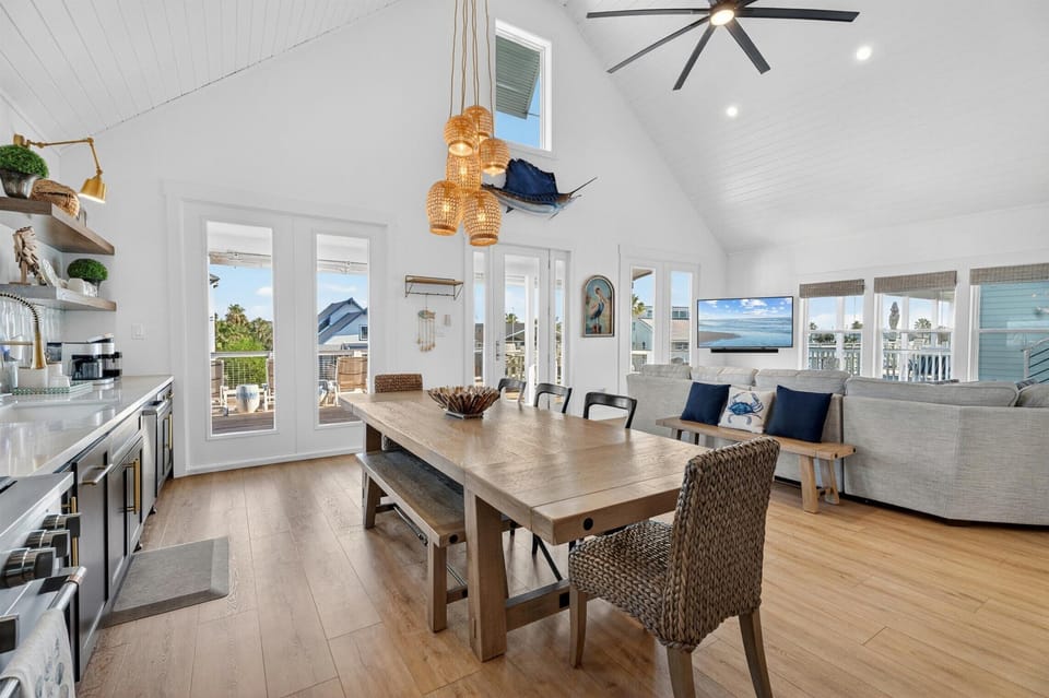 Open dining area with large table, pendant lights, TV lounge, and bright deck doors.