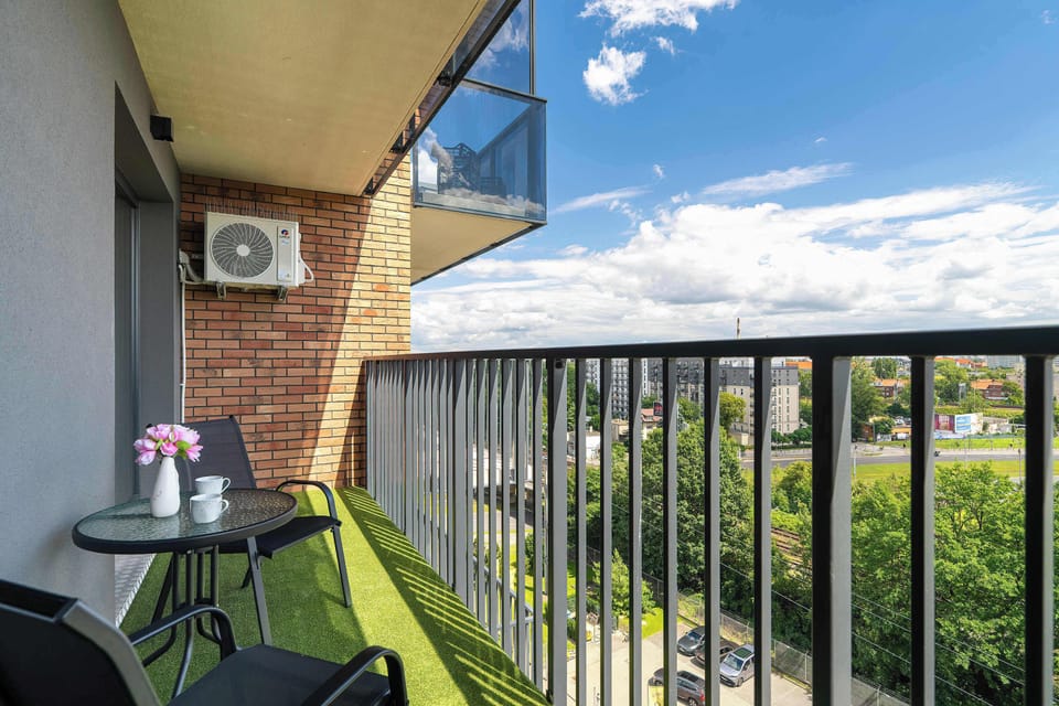 A compact balcony covered with green artificial turf, black metal railings, and a small bistro table set. Sunny outdoor views and neighboring greenery surround the space.