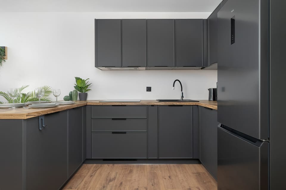 A contemporary kitchen space showcases sleek dark grey lower cabinetry topped with light wooden countertops. A tall black refrigerator stands at the end of the counter line under bright overhead lighting.