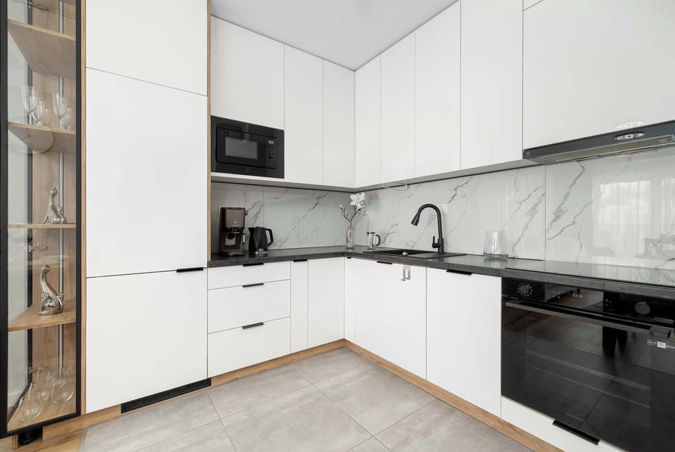 A corner-angled view of the L-shaped white kitchenette highlights the ample counter space and dark tiled backsplash. Modern appliances, including an oven and a coffee machine, are seamlessly built into the layout.