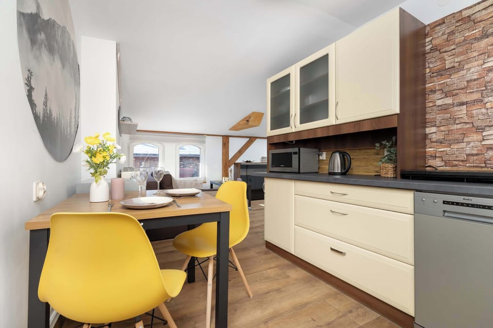 A compact dining area with vibrant yellow chairs connects to a modern kitchenette. Brick-patterned wall tiles create a stylish contrast against light cabinets.
