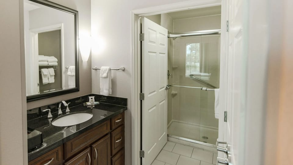 Bright bathroom features a dark wood vanity, granite countertop, modern mirror, and glass shower with white tiles and fluffy towels