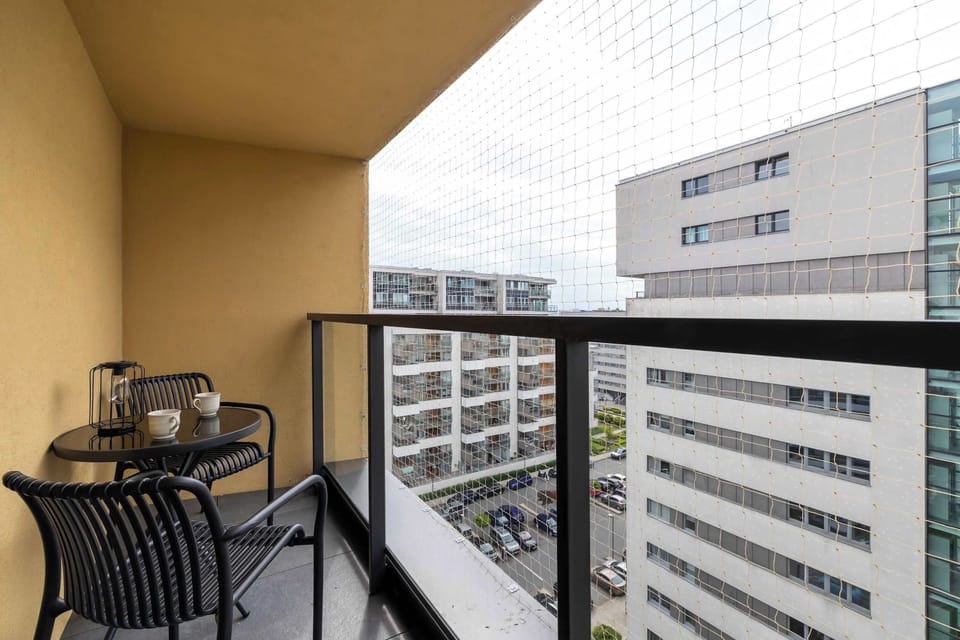 A cozy private balcony furnished with a small table and black chairs. High-rise city buildings are visible in the background under daylight.