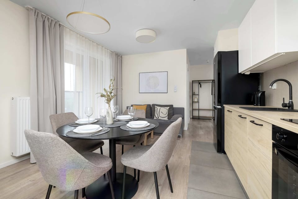 A wide view of an apartment showing a dining space connected to a modern kitchen kitchenette. The room features neutral tones and stylish decor.