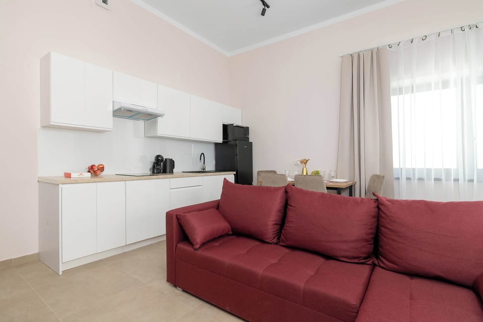 An open-plan room highlights a white modern kitchen counter along the left wall and a large sofa on the right. Sheer curtains cover the far window, illuminating the spacious layout.