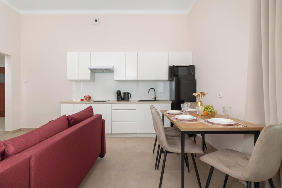 A dining space with a light wooden table and gray padded chairs is positioned near a white kitchenette setup and a sofa. Light wall finishes keep the atmosphere bright and open.
