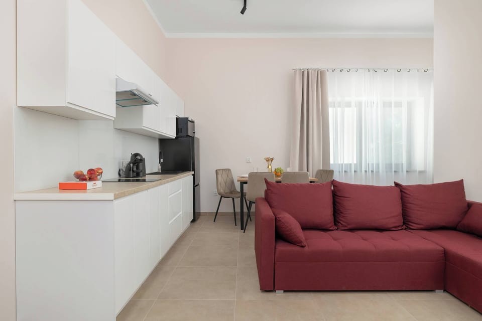 A bright kitchenette and living area showcases a sleek white countertop with built-in appliances and a red sectional sofa in the background. Large windows let in plenty of natural light.
