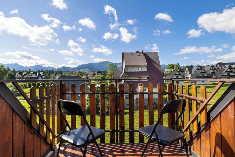 A wooden balcony with two chairs overlooks green fields and traditional mountain architecture under a cloud-filled sky.
